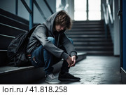 Lonely upset teen guy in hooded jumper with backpack sitting on step of the stairs, adolescence problems. Стоковое AI-изображение, AI-художник Оксана Гильман / Фотобанк Лори