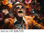 Portrait of overjoyed old eccentric senior man in orange suit dancing in front of a colorful flowers background. Expressing joy and free spirit, beauty of aging and individuality and carelessness. Стоковое AI-изображение, AI-художник Ирина Михайличенко / Фотобанк Лори