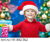 Smiling boy in a red Christmas hat with gifts near the Christmas tree. Стоковое AI-изображение, AI-художник Сергей Елизарьев / Фотобанк Лори