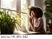 Happy young dark skinned woman sitting with laptop and smartphone mear window with green plants. Стоковое AI-изображение, AI-художник Наталья Семкина / Фотобанк Лори
