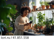 Dark skinned young woman holding smartphone in his hands, studying information about house plants. Стоковое AI-изображение, AI-художник Наталья Семкина / Фотобанк Лори