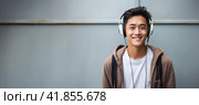 Cheerful asian young man listening music with headphones on grunge gray wall background. Copy space. Стоковое AI-изображение, AI-художник Наталья Семкина / Фотобанк Лори
