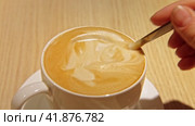 A Hand is Mixing the Milk Foam of Latte. Person Stirring Cappuccino with Steel Spoon. Hot Coffee in a White Ceramic Cup on a Wooden Table. Modern Coffee shop, Cafeteria. Thick Froth. Close-up View. Стоковое видео, видеограф Марина Демидюк / Фотобанк Лори