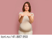 A pregnant woman shows her palms with a plea for help on a studio pink background. Pregnancy in a woman with a belly. Стоковое фото, фотограф Андрей Журавлев / Фотобанк Лори