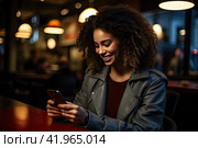 An African girl is sitting at a table in a cafe and smiling at the screen of her smartphone. The concept of communication at leisure. Стоковое фото, фотограф Ярославский Максим / Фотобанк Лори