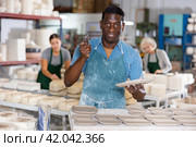 Portrait of african american worker cutting burrs off freshly made earthenware cups and plates with knife. Стоковое фото, фотограф Яков Филимонов / Фотобанк Лори
