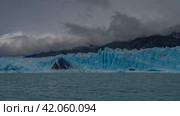 A wall of blue ice with cracks and crevices in a glacial lake. A dark triangular iceberg has broken off, melted ice floes float in turquoise water. The mountains are in a fog. Perito Moreno glacier. Стоковое фото, фотограф Вера Ивановна Кубарева / Фотобанк Лори
