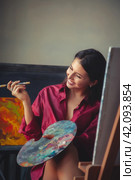 Lovely lady art painter with palette and brush in red shirt sitting at canvas, laughing. Happy perfect drawing woman artist painting in home art lab. Artisanal lifestyle concept. Copy ad text space. Стоковое фото, фотограф Aleksandr Golubev / Фотобанк Лори