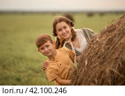 Дети, девочка и мальчик, выглядывают из-за стога сена и улыбаются. Стоковое фото, фотограф Анастасия Пипко / Фотобанк Лори