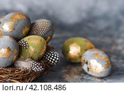 Stylish beautiful Easter eggs with a golden coating of sweat and feathers in a bird's nest of brushwood on a gray background. The concept of happy Easter 2024. Стоковое фото, фотограф Photojuli / Фотобанк Лори
