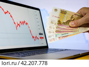 Vladimir, Russia - February 2022: Paper banknotes of Russian money in a male hand against the background of declining financial charts. The concept of crisis and devaluation. Selective focus on banknotes. Редакционное фото, фотограф Никита Бурденков / Фотобанк Лори