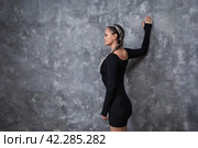 A brunette woman leading a healthy lifestyle performs an exercise to reduce stoop, trains in sports black clothes, leaning with one hand on the wall and placing it behind her back, with copy space. Стоковое фото, фотограф Сергей Чайко / Фотобанк Лори