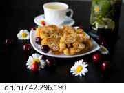 На белой тарелке десерт из ирисок и кукурузных палочек, украшенный ромашками с божьими коровками и вишнями. Рядом чашка травяного чая и чайник, в котором заварены лимон, мята и ромашка. Стоковое фото, фотограф Елена Яворская / Фотобанк Лори