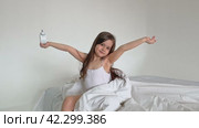 A nice child girl enjoys sunny morning. Good morning at home. Child girl wakes up from sleep. Sweet little girl is looking at camera and smiling while sitting on her bed on white background. Стоковое видео, видеограф Ирина Михайличенко / Фотобанк Лори