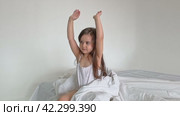 A nice child girl enjoys sunny morning. Good morning at home. Child girl wakes up from sleep. Sweet little girl is looking at camera and smiling while sitting on her bed on white background. Стоковое видео, видеограф Ирина Михайличенко / Фотобанк Лори