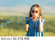 Portrait of funny lovely little girl in blue drees and sunglasses blowing soap bubbles on nature background. Happy childhood and summer concept. Стоковое фото, фотограф Ирина Михайличенко / Фотобанк Лори