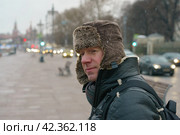 Handsome man walking in Moscow city center. Cold winter in the Russian capital during coronavirus pandemic time. He wear hot cap made of natural sheepskin. It fastened at the chin to protect from cold. Стоковое фото, фотограф YuryKara / Фотобанк Лори