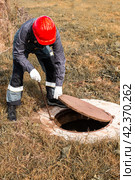 A worker in a hard hat lifts a manhole cover on a septic well. Inspection and maintenance of sewerage systems in rural areas. Стоковое фото, фотограф Коханова Наталья / Фотобанк Лори
