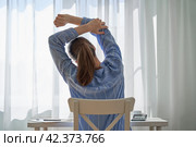 Back view of young faceless woman in casual clothes stretching. Remote work challenges and disadvantages. Working at home difficulties. Downsides of hardworking. Quarantine workspace. Neutral tones. Стоковое фото, фотограф Natalia Gdovskaia / Фотобанк Лори