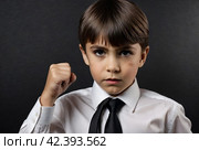 Serious boy in shirt and tie shows fist to revenge or threatening with angry look. Annoyed face boss, looking at camera. Evil teen child on isolate black background. Child emotion concept. Copy space. Стоковое AI-изображение, AI-художник Aleksandr Golubev / Фотобанк Лори