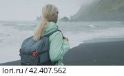 Woman with a green raincoat walking on a black sand beach enjoying view to Reynisfjara sea stack cliff formations close to Vik, Iceland. Стоковое видео, видеограф Igor Tichonow / Фотобанк Лори