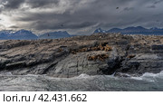 A family of sea lions is resting, lying on the slope of a rocky islet. Cormorants are sitting on cliffs, flying in the cloudy sky. Mountains in the distance. The waves are foaming. Isla de los lobos. Стоковое фото, фотограф Вера Ивановна Кубарева / Фотобанк Лори