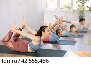 Middle-aged woman does yoga or Pilates-Dhanurasana, Bow pose. Family in background. Стоковое фото, фотограф Яков Филимонов / Фотобанк Лори