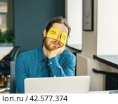 Tired sleeping young male office worker hiding eyes with funny sticky notes. Стоковое фото, фотограф Natalia Gdovskaia / Фотобанк Лори