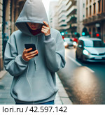 Подросток в худи с капюшоном на улице города залипает в соцсетях в смартфоне. Опасная зависимость от гаджетов и невнимательность. Номофобия - страх остаться без телефона. Стоковое AI-изображение, AI-художник Sergei Gorin / Фотобанк Лори