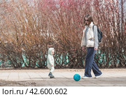 A handsome, cheerful little boy of two years old walks on the street in the spring. A kid in a jumpsuit plays ball with his mother. The concept of parenthood, childhood and family. Стоковое фото, фотограф Photojuli / Фотобанк Лори