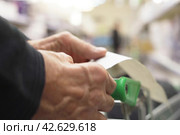 The hands of an unrecognizable older man hold a sales receipt next to a grocery cart in a supermarket. Стоковое фото, фотограф Владимир Федоров / Фотобанк Лори