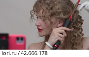 A blonde blogger creates a stunning new look by manually wrapping strands of long hair around a curling iron while live streaming to social media on her smartphone (2024 год). Редакционное видео, видеограф А. А. Пирагис / Фотобанк Лори