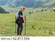 Rear view of hiker female with backpack and trekking pole standing on green lawn on the backdrop of forest and watching at camera. Girl going trek by outdoor spaces. Concept of travelling, hiking. Стоковое фото, фотограф Ирина Михайличенко / Фотобанк Лори