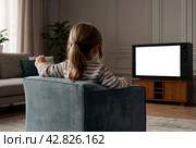 Rear view of little girl kid 5-6 year old watching tv sits on armchair looking cartoon. Adorable dependency girl child, empty isolated screen. Television addiction tv concept. Copy ad text space. Стоковое AI-изображение, AI-художник Aleksandr Golubev / Фотобанк Лори