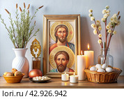 Orthodox Easter. Easter cake, painted eggs, icons, faces of saints, flowers, church candles and Easter decor on a wooden table in an old village house. Стоковое AI-изображение, AI-художник Photojuli / Фотобанк Лори