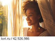 Young african american woman unveil curtain and looking out of window in the morning. Beautiful girl after waking up fully rested and open curtains. Start the new day with happiness. Relax, resting. Стоковое AI-изображение, AI-художник Марина Демидюк / Фотобанк Лори