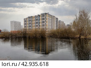 Жилые дома района новостройки отражаются в воде маленького озера, окруженного кустами и деревьями. Пасмурное небо с облаками, через которые пробиваются солнечные лучи. (2024 год). Редакционное фото, фотограф Сергей Никифоров-Гордеев / Фотобанк Лори