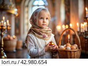 Happy bright Easter. Christianity. Portrait of a three-year-old girl in a Russian folk dress and shawl with a wicker basket with Easter eggs in her hands against the background of an Orthodox church. Стоковое AI-изображение, AI-художник Photojuli / Фотобанк Лори