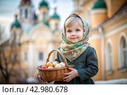 Happy bright Easter. Christianity. Portrait of a three-year-old girl in a Russian folk dress and shawl with a wicker basket with Easter eggs in her hands against the background of an Orthodox church. Стоковое AI-изображение, AI-художник Photojuli / Фотобанк Лори