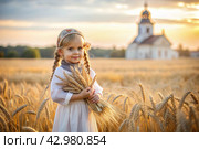 Russian Russian girl in a white dress and a Russian headscarf holds golden ears of wheat against the background of a ripening field and an Orthodox church. The concept of planting and harvesting a rich harvest. Rural landscape. Стоковое AI-изображение, AI-художник Photojuli / Фотобанк Лори