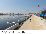Набережная реки Амур в российском городе Благовещенске на границе с Китаем. Амурская область, Дальний Восток России. По набережной на электрическом самокате едет юноша с девушкой. На флагштоке флаг России. Вдали на противоположном берегу реки виден китайский город Хэйхэ. На воде льдины. Весенний сезон. Апрель (2023 год). Редакционное фото, фотограф Андрей Степанов / Фотобанк Лори