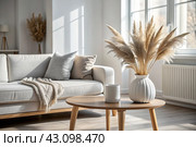 A modern white ceramic vase with dried grass on a wooden table. Scandinavian interior. An empty white wall with a shadow from the window. Стоковое AI-изображение, AI-художник Photojuli / Фотобанк Лори