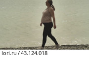 Беременная женщина безмятежно прогуливается по галечному берегу моря. Она одета в повседневную одежду. Спокойная вода рядом отражает безмятежную атмосферу. Стоковое видео, видеограф Воловодов Владимир / Фотобанк Лори
