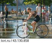 Moscow, Russia - July 3, 2022: Young woman cycling through the water of fountain in sunny summer day. He is having fun. Boy rides bike. Hot summer amusement. Leisure concept. Side view. Редакционное фото, фотограф YuryKara / Фотобанк Лори