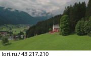 Serene hillside view featuring a small pink chapel and lush green surroundings, with a distant town and forested mountains. Стоковое видео, видеограф Igor Tichonow / Фотобанк Лори