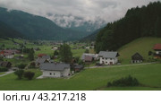 Serene village with houses scattered on a green hillside, surrounded by forested mountains and a cloudy sky, creating a peaceful atmosphere. Стоковое видео, видеограф Igor Tichonow / Фотобанк Лори