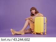 A thoughtful traveler girl is sitting next to a yellow suitcase on a uniform lilac background. Copy space. Стоковое AI-изображение, AI-художник Ярославский Максим / Фотобанк Лори