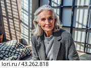 Minimalism. A studio portrait of a stylish, elegant elderly woman, a pensioner of 70 years old, in an interior and clothes of calm tones. The concept of the elderly. Стоковое AI-изображение, AI-художник Photojuli / Фотобанк Лори