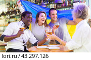Cheerful diverse soccer supporters holding the flag of Ukraine and enjoing beer, spending time together in the sport bar. Стоковое фото, фотограф Яков Филимонов / Фотобанк Лори