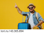 A male traveler and a blue suitcase on a yellow background shows his hand up. Copy space. Стоковое AI-изображение, AI-художник Ярославский Максим / Фотобанк Лори